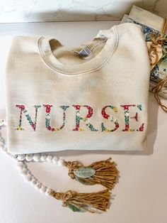 This embroidered floral font is absolutely beautiful. The detail to the flowers and colors are stunning.  Nurse embroidered to the front. Approximately 11x2 inches. Spring White Sweatshirt With Custom Embroidery, White Custom Embroidered Sweatshirt For Spring, White Sweatshirt With Custom Embroidery For Spring, Embroidered Long Sleeve Sweatshirt For Spring, Cream Cotton Sweatshirt With Letter Embroidery, White Floral Embroidery Sweatshirt For Spring, White Floral Embroidered Sweatshirt For Fall, Spring Cream Cotton Sweatshirt, Fall Cotton Sweatshirt With Floral Embroidery