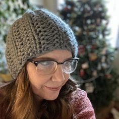 a woman wearing glasses and a knitted hat looks at her cell phone while sitting in front of a christmas tree