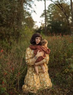 a woman holding a child in her arms while standing in the grass with trees and bushes behind her