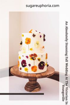 a three layer white cake with flowers on it sitting on top of a wooden stand