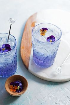 two glasses filled with blue liquid and purple pansies on top of a wooden tray