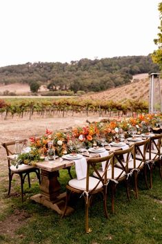 an outdoor dining table set up with flowers and greenery in front of a vineyard