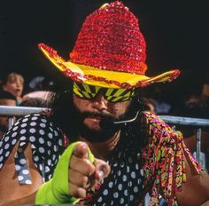 a man wearing a colorful hat and sunglasses pointing to the side while sitting in front of a crowd