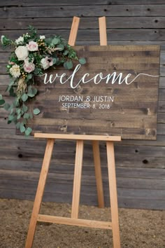 a wooden welcome sign with greenery and flowers on it, sitting in front of a barn