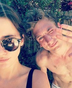 two people standing next to each other in front of a wooden fence and green plants