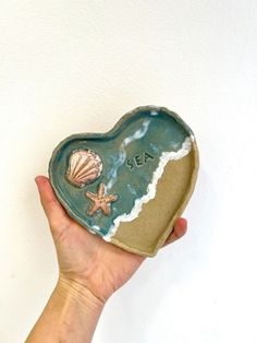 a hand holding a ceramic heart shaped dish with seashells and starfish on it