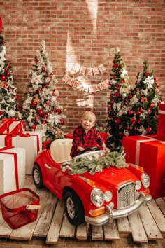 a little boy sitting in a red toy car surrounded by christmas trees and wrapped presents