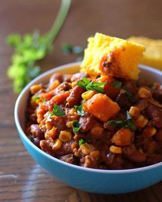 a blue bowl filled with chili and corn next to a piece of bread on top of a wooden table