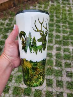 a hand holding a coffee cup with trees and deer on it, in the grass