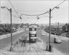 a black and white photo of a train on the tracks in an old town with cars driving by