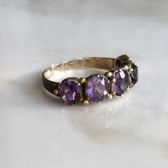 A late Georgian (circa.1830-1837) 15 carat gold Amethyst ring. This beautiful piece holds all five stones neatly in line, what a gorgeous collection of glowing purple stones. The shoulders show a nice bit of detailing too. CONDITION: Wear consistent with age and use. Centre stone previously replaced. Please see photos for more detail. SETTING (ACROSS): 30mm STONE DIAMETER: 5mm BAND WIDTH (SMALLEST): 2mm RING SIZE: UK: N 1/2 | US: 6 3/4 WEIGHT: 2.4 grams (SZZ) White Stone Ring, Gold Amethyst Ring, Amethyst Gem, Purple Band, Purple Stones, Delicate Rings, Lovely Jewellery, Multi Stone Ring, Gold Platinum