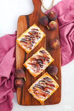 three pieces of bread on a wooden cutting board with figs next to it and a pink napkin