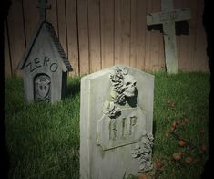 two tombstones sitting in the grass next to a wooden fence and building with a skull on it