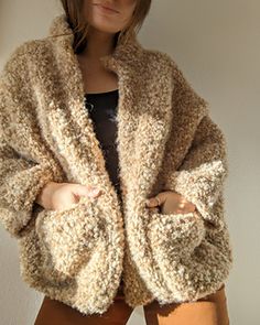 a woman standing in front of a white wall wearing a brown coat and tan pants