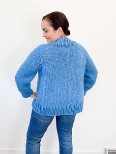 a woman standing in front of a white wall wearing a blue sweater