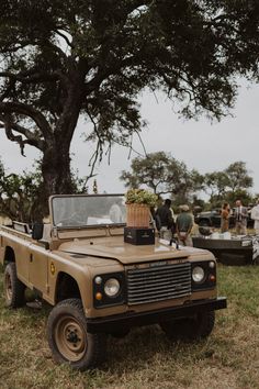 an old jeep is parked in the grass with people standing around it and some trees