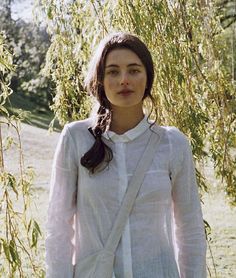 a woman standing under a tree with her hands in her pockets and looking at the camera