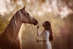 a woman standing next to a brown horse in the woods with her hand on it's bridle