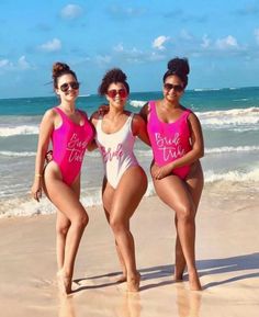 three women in pink and white swimsuits standing on the beach with their arms around each other