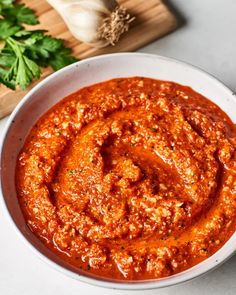 a white bowl filled with tomato sauce next to some parsley on a cutting board