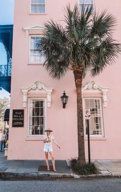 a woman standing in front of a pink building with a palm tree next to it