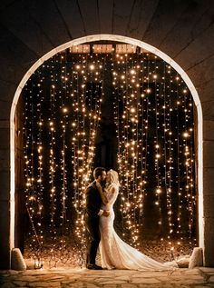 a bride and groom standing in front of an archway covered with fairy lights