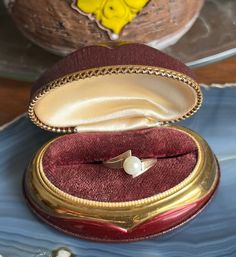 an open ring box with a pearl in it sitting on a blue plate next to a yellow flower