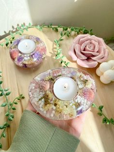 a hand holding a glass candle holder with flowers and leaves around it on a table