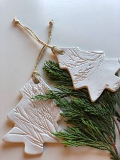 three white ceramic ornaments hanging from twine with green leaves and pine cones on them