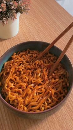 a bowl filled with noodles and chopsticks on top of a wooden table next to a potted plant