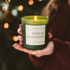 a woman holding a candle in front of a christmas tree with the words home for the holidays written on it