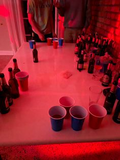 there are many cups on the table with beer bottles around it and one person is standing in the background