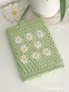 a green crocheted notebook with white flowers on it next to a flower pot