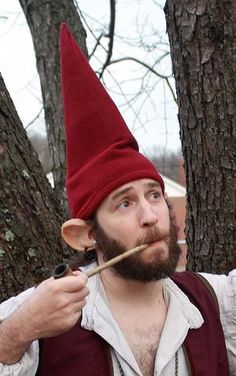 a man wearing a red hat and holding a stick in his mouth while standing next to a tree