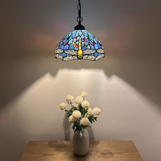 a vase with white flowers on a table under a stained - glass light fixture, next to a lamp shade