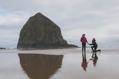 two people holding hands on the beach near a large rock in the distance with water reflecting off it's surface