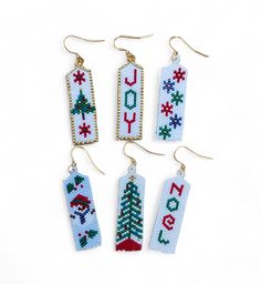 cross stitch christmas ornaments hanging from gold earwires on a white background with the words joy written in red, green and blue