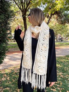 a woman standing in the grass wearing a white crochet scarf with tassels