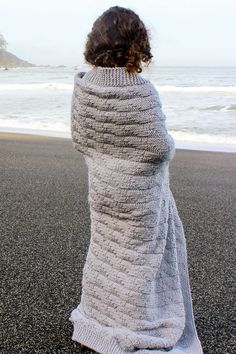 a woman standing on the beach wearing a knitted shawl and looking out at the ocean