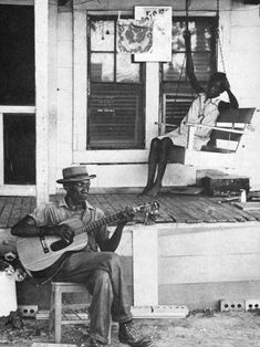 black and white photograph of man playing guitar on porch with woman sitting in window behind him