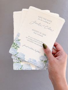 a hand holding three wedding cards with blue flowers on the front and green leaves on the back