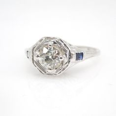 When you picture an "Art Deco" engagement ring, one or two different visions probably pop into your head. At least one of those visions surely resembles this ring.At the center of this 18K white gold ring is a 5mm European Cut diamond (weighing at least half a carat), held in place with four prongs and set into a floating plate gently separated from the rest of the ring. Underneath the center stone is a beautifully crisp filigree pattern reminiscent of a sunrise... and two deep baguette sapphire Octagon Brilliant Cut Sapphire Ring With Diamond, Octagon White Gold Sapphire Ring With Diamond Cut, Formal Octagon Sapphire Ring With Diamond Cut, White Gold Octagon Sapphire Ring With Brilliant Cut, Formal Sapphire Ring With Octagon Diamond Cut, White Gold Sapphire Ring With Brilliant Octagon Cut, Formal Octagon Brilliant Cut Sapphire Ring, Brilliant Cut Octagon Sapphire Ring In Platinum, Formal Octagon Sapphire Ring With Brilliant Cut