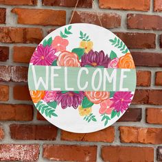 a welcome sign hanging on the side of a brick wall with flowers painted on it