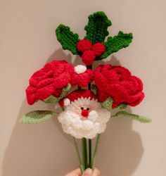 a hand holding a crocheted santa clause bouquet with red flowers and green leaves