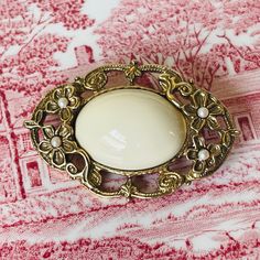 Vintage 1928 Cream Oval Cabochon & Seed Pearl Gold Brooch (1970s) Description: Embrace the timeless elegance of this vintage 1928 brooch from the 1970s. This exquisite piece features a large cream-colored oval cabochon set in a beautifully detailed gold-tone frame, accented with delicate floral motifs. Each flower is adorned with a small seed pearl, adding a touch of sophistication to the design. The back of the brooch is imprinted with the signature hallmark pattern of the 1928 jewelry company, Vintage 1928 Jewelry, Antique Oval Cabochon Brooch For Formal Occasions, Classic Oval Cabochon Brooches, Antique Gold Brooch With Oval Cabochon, Antique Gold Brooches With Oval Cabochon, Antique Gold Oval Cabochon Brooch, Antique Gold Oval Cabochon Brooches, White Vintage Cabochon Brooches, Classic Gold Oval Brooch