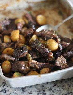 a bowl filled with meat and potatoes sitting on top of a marble counter next to a spoon