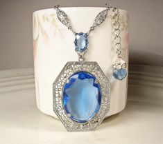 Offering an absolutely stunning vintage 1920s to 1930s era Art Deco sapphire blue bezel set faceted glass crystal and silver rhodium plated filigree pendant necklace.  This is the perfect "Something Old" and "Something Blue" for the bride!   The 1 5/8 inch (41 cm) by 1 1/4 inches at 1 5/16 inches (3.3 cm) ornate embossed filigree pendant features a central oval cut bezel set faceted glass crystal in the most beautiful shade of sapphire blue.  It hangs from a smaller open back faceted glass crystal also in blue.  This in turn is suspended from rhodium plated filigree links on either side.  The rest of the necklace is a platinum tone replacement chain with that fastens with a ring clasp.  It has an extender chain for versatility and can be worn from about and measures 21 3/8 to 23 7/8 inches Art Deco Pendant Necklace, Pendant Necklace Long, Blue Sapphire Jewelry, Filigree Pendant Necklace, Vintage Rhinestone Jewelry, 1920s Vintage, Art Deco Pendant, Filigree Pendant, Chain Fashion