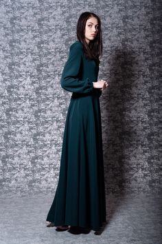 a woman in a long green dress standing against a gray wall with her hands on her hips