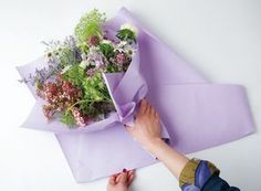 someone wrapping flowers in purple paper on top of a white surface with one hand reaching for the flower