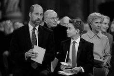 two men sitting next to each other in suits and ties with people standing behind them